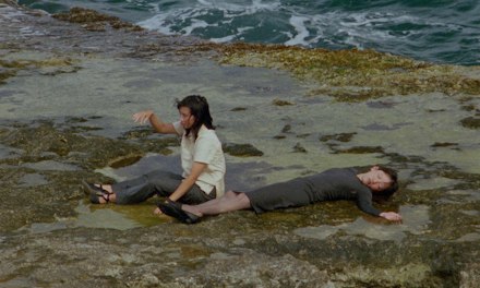 A colour photograph of two people lying in shallow seawater among rocks and seaweed.